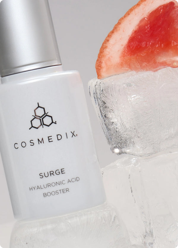Close up of Surge Hyaluronic Acid Booster next to ice block with grapefruit  against light gray background