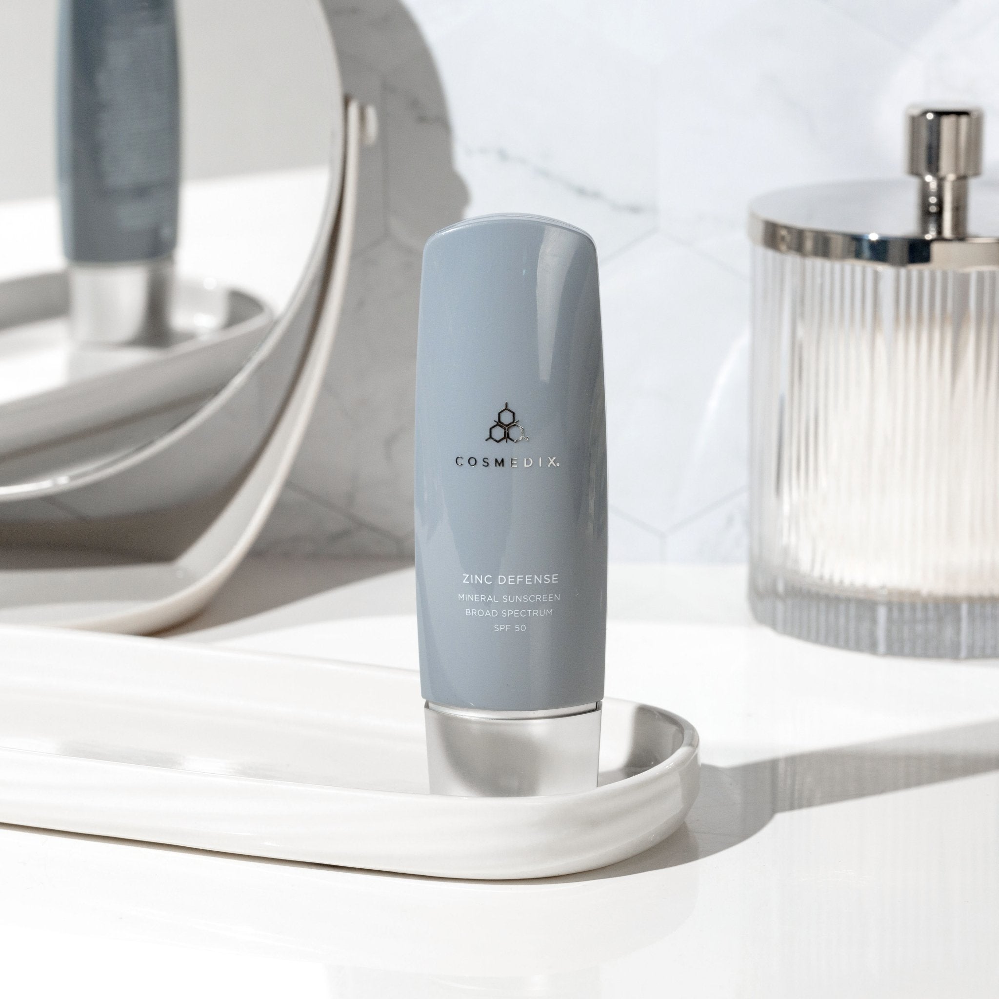 CosMedix Zinc Defense mineral sunscreen bottle on a white marble counter with a pump bottle in the background.